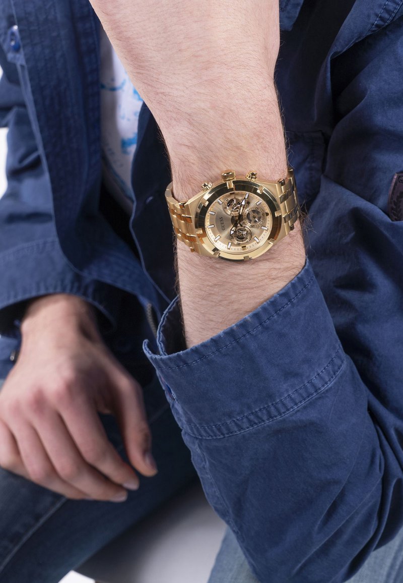 Montre en or avec un bracelet métallique, cadran rond, cadran transparent montrant les engrenages, et trois sous-cadrans. Portée au poignet, assortie à une chemise bleue.