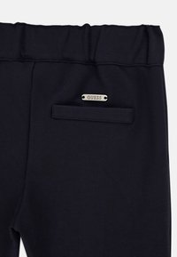 Dos d'un pantalon noir avec une taille élastique, une poche passepoilée unique et une petite étiquette rectangulaire argentée portant l'inscription "GUESS" au-dessus de la poche.