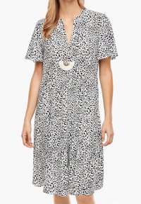 Kortärmad, lager-på-lager klänning i vitt tyg med svarta leopardfläckar. Har en V-ringning och ett stort, runt hängsmycke.