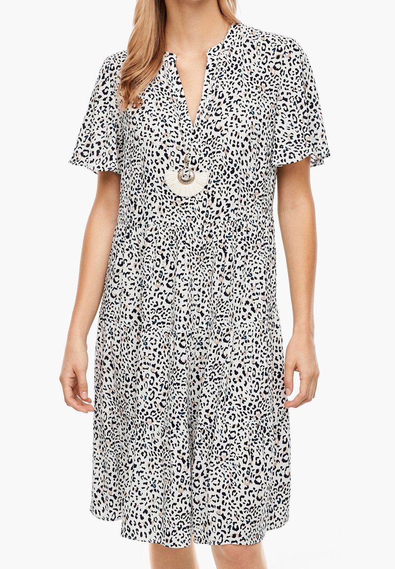 Kortärmad, lager-på-lager klänning i vitt tyg med svarta leopardfläckar. Har en V-ringning och ett stort, runt hängsmycke.