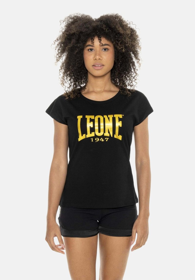 Femme aux cheveux bouclés portant un t-shirt noir avec un texte doré "LEONE 1947" et un short noir, debout sur un fond blanc.