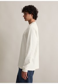 Langärmliges weißes Baumwoll-T-Shirt mit Rundhalsausschnitt, ausgestattet mit gerippten Bündchen und einem lässigen Schnitt, kombiniert mit blauen Denim-Hosen.