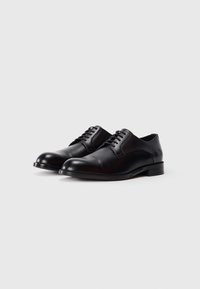 Un paio di scarpe eleganti in pelle nera, con lacci, punte arrotondate e tacchi bassi, su uno sfondo chiaro e semplice.