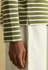 Langärmliges Oberteil mit olivgrünen und weißen Streifen und abgerundetem Saum, kombiniert mit einem weißen, strukturierten Stoffstück. Sichtbare Hand ruht auf dem Stoff.