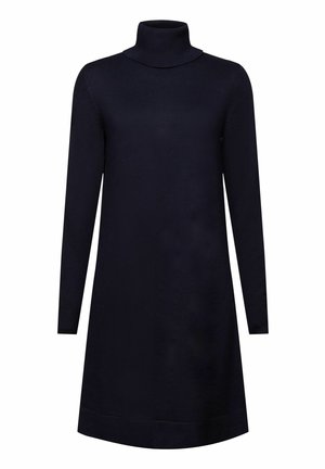 Femme aux longues tresses portant une robe bleu marine, un pull vert clair noué autour de la taille, des chaussettes noires et des bottines beiges, debout de dos.