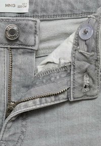 Sive jeans iz denima imajo zadrgo, gumbasto zapiranje in vidne šive. Notranja podloga je bela, z raztrganimi robovi v bližini zadrge.