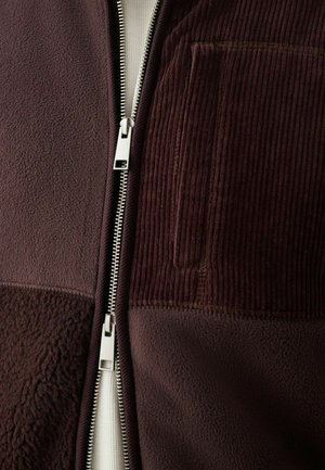 Burgundi színű, cipzáras dzseki, amely bolyhos és kordbársony textúrával rendelkezik, ezüst cipzárral és egy kis mellzsebbel. A sima bélés látható.