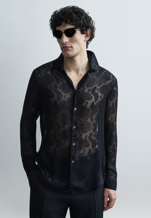 Muž oblečený v čiernej priesvitnej vzorovanej košeli na gombíky, čiernych nohaviciach a čiernych slnečných okuliaroch, stojaci pred svetlosivým pozadím.