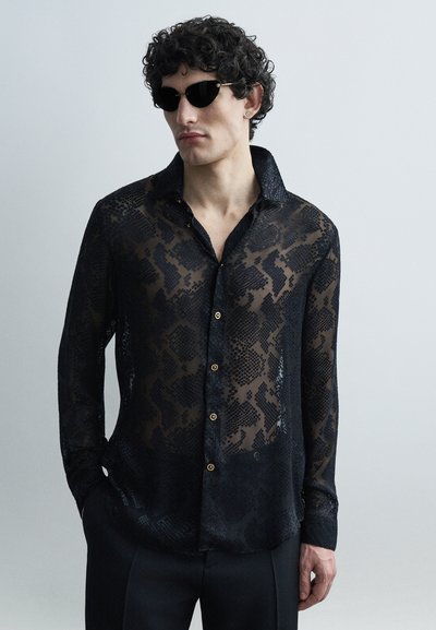 Homme portant une chemise noire transparente à motifs avec des boutons, un pantalon noir et des lunettes de soleil noires, debout devant un fond gris clair.