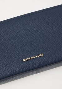 Tumši zila ādas maku ar teksturētu virsmu un zelta "MICHAEL KORS" logo. Iezīmējas ar elegantu dizainu ar minimālu šuvju un akcentu skaitu.