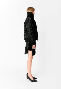 Chaqueta estructurada negra con diseño de cintas en capas, combinada con una falda que presenta acentos a juego. Llevada con tacones negros puntiagudos.