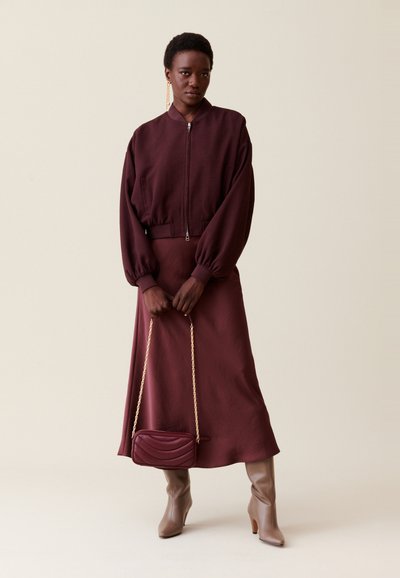 Veste bomber bordeaux avec des manches bouffantes, jupe assortie bordeaux, bottes brunes jusqu'aux genoux, et un petit sac à bandoulière matelassé avec une chaîne dorée.