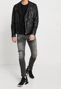 Chaqueta de motocicleta de cuero negro con cremalleras, usada sobre una camiseta negra; combinada con unos jeans grises desgastados y zapatillas deportivas negras.