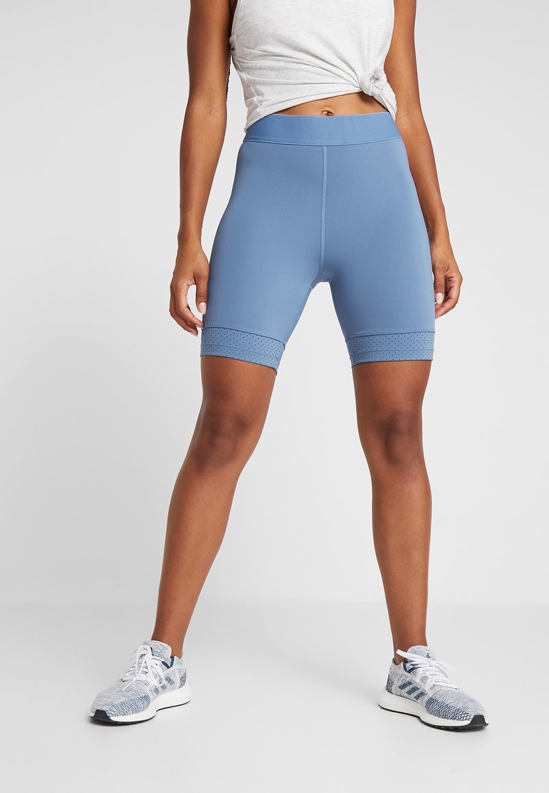 Femme portant un short de sport ajusté bleu clair et des chaussures de course blanches à motifs, debout devant un fond clair uni.