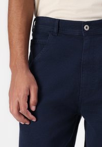 Navy blue denim pants with a straight cut, featuring a zip fly, front pockets, and a textured surface. Metal button closure visible.