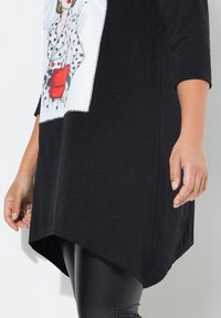 Schwarze Tunika mit einem weißen Paneel, das eine Polka-Dot-Illustration einer Frau zeigt. Der Saum ist hinten länger und hat eine glatte, weiche Stofftextur.