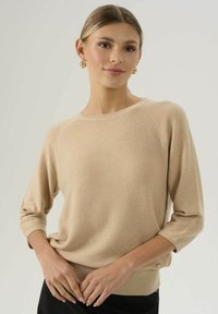 Beżowy sweter dzianinowy z rękawami trzyczwartowymi, okrągłym dekoltem, prążkowanym wykończeniem i ozdobnymi guzikami z boku. Gładka tekstura.
