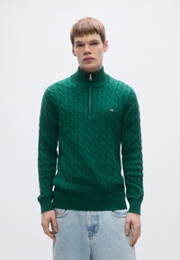 Zelený pletený sveter s vysokým golierom, so zipsom a vzorom káblového pletenia. Rebrované manžety a lem, nosený cez svetlomodré rifle.