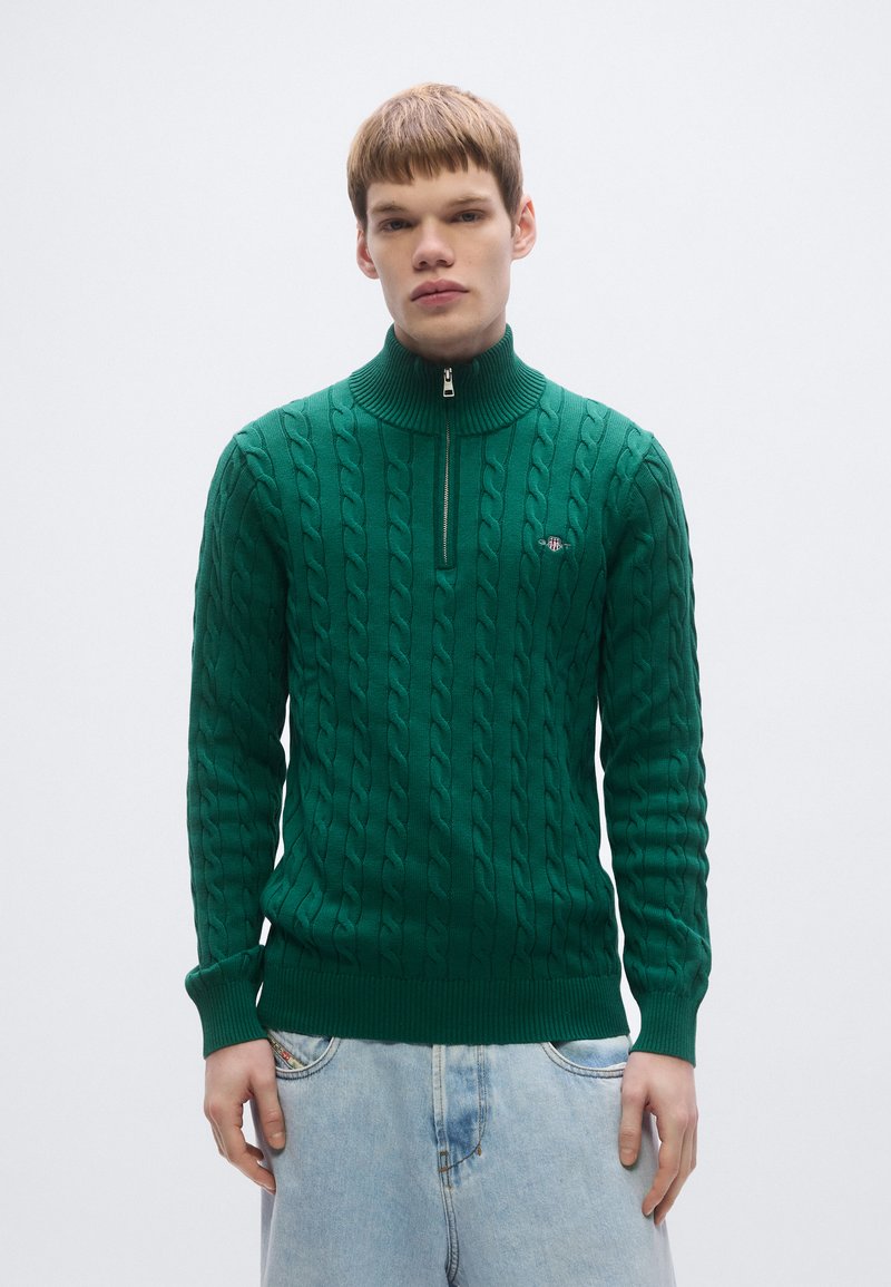 Zelený pletený sveter s vysokým golierom, so zipsom a vzorom káblového pletenia. Rebrované manžety a lem, nosený cez svetlomodré rifle.
