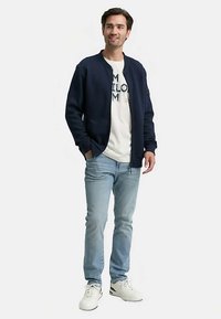 Marineblaue Bomberjacke mit gerippten Bündchen, getragen über einem hellgrauen T-Shirt mit Grafik. Hellblaue Jeans und weiße Sneaker vervollständigen das Outfit.