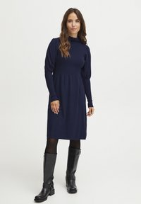 Marinblå stickad klänning med långa ärmar och hög krage, med en figursydd överdel och en utsvängd kjol, i kombination med knähöga svarta stövlar.