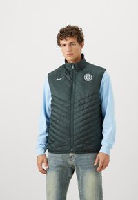 Nike kviltet vest i mørkegrøn, med lynlås foran, høj krave, Chelsea FC-logo på brystet, båret over en lyseblå langærmet skjorte.