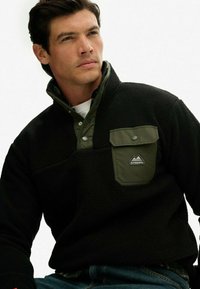 Sort fleece pullover med en grøn knapkrave, der har en brystlomme og et logo patch. Blød tekstur og afslappet pasform.