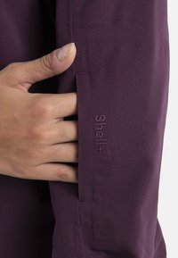 Fioletowy materiał kurtki o gładkiej fakturze, z logo 'Shell+' wyhaftowanym w dopasowanym odcieniu na rękawie. Ręka chwytająca materiał.