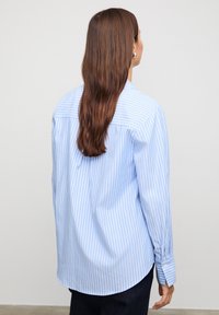 Blue and white striped button-up shirt with long sleeves, curved hem, and a back yoke detailing. Fabric appears smooth and tailored.
