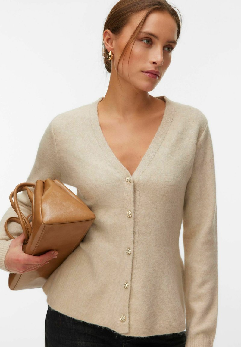Femme portant un cardigan beige boutonné et des boucles d'oreilles dorées, tenant un sac à main en cuir couleur tabac devant un fond blanc uni.