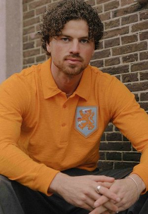 Oranje polo met lange mouwen en een blauw schildpatch met een leeuw. De stof lijkt zacht met een gestructureerde afwerking; kraag met knopen.