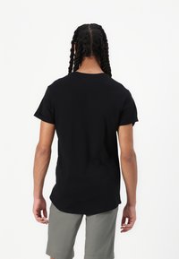 Tricou negru cu mânecă scurtă, cu guler rotund și croială lejeră, asortat cu șorturi gri deschis, văzut din spate.