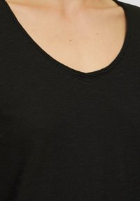 Juodas V-kaklo marškinėlis su tekstūruota medžiaga, turintis paprastą siūlę aplink kaklą ir laisvą siluetą. Lygus paviršius su subtiliu blizgesiu.