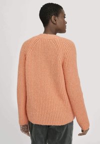 Person mit kurzen Haaren, die einen strukturierten, pfirsichfarbenen Strickpullover und dunkle Hosen trägt, von hinten vor einem schlichten Hintergrund gezeigt.