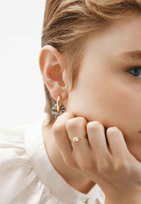 Arete de oro con un colgante circular a lunares. Superficie lisa que resalta un contraste de materiales. El modelo tiene el cabello corto y un anillo en el dedo.