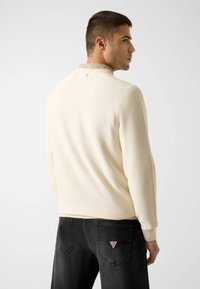 Maglione color crema con finitura testurizzata, caratterizzato da un colletto alto e polsini a costine, abbinato a jeans di denim neri con un logo a triangolo.