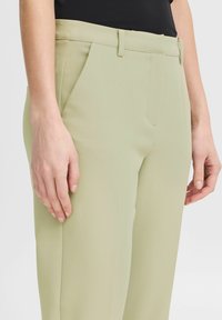 Svetlozelene krojene hlače z gladko teksturo, ki imajo ravno sprednjo stran, zanke za pas in stranske žepe, delno prikazane s roko.