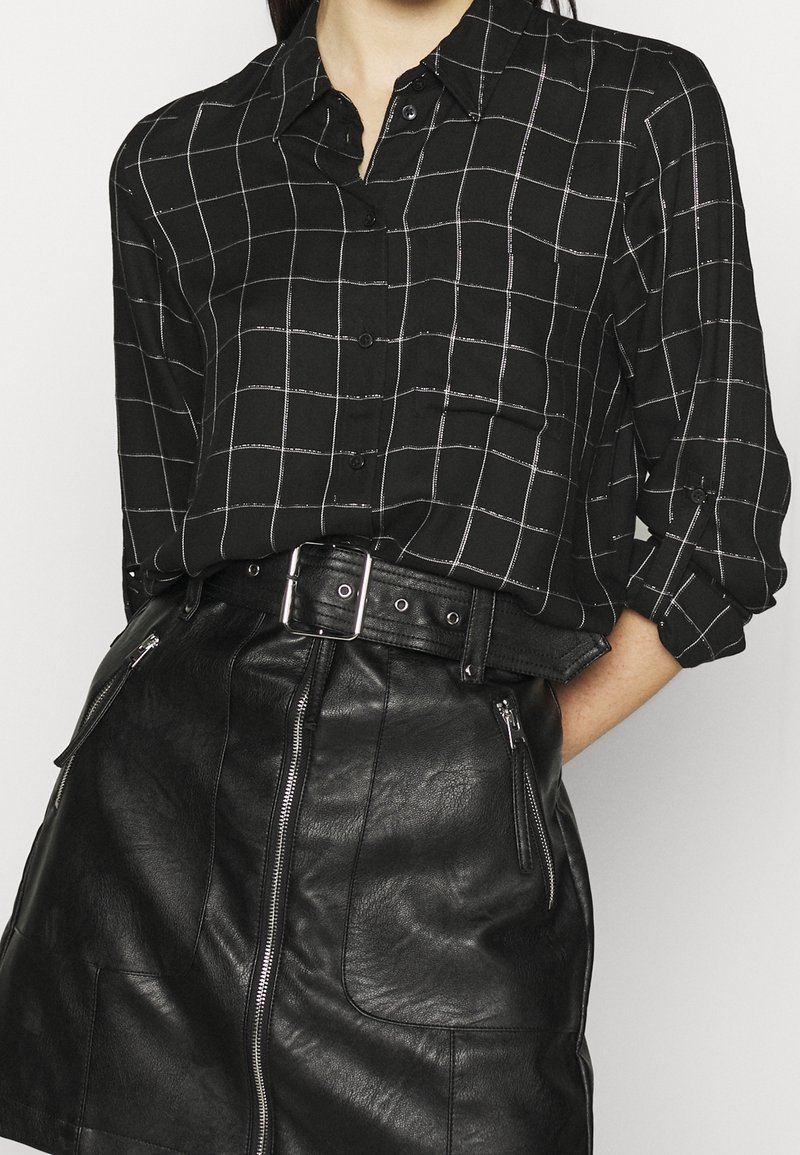 Femme portant une chemise à carreaux noire boutonnée, rentrée dans une jupe en cuir noire avec des poches zippées et une ceinture avec une boucle argentée.