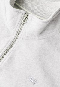 Sudadera gris con cuello con cremallera y textura acanalada. Presenta un pequeño logo bordado en la esquina inferior. Tela suave.