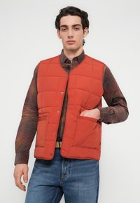 Κόκκινο γιλέκο με ραφές σε σχήμα καπιτονέ, με κουμπιά-κλίπ και πλαϊνές τσέπες, φορεμένο πάνω από ένα καφέ καρό πουκάμισο. Συνδυάζεται με μπλε τζιν και ανοιχτόχρωμη ζώνη.