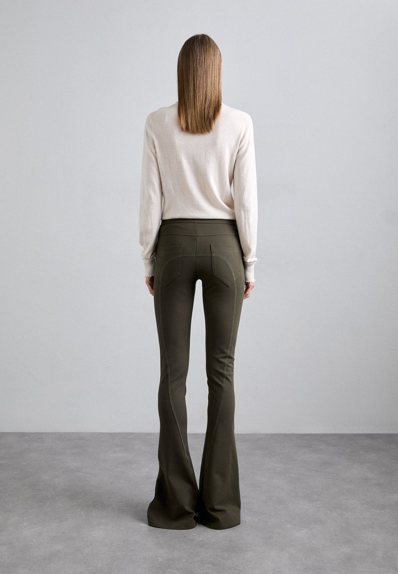 Olive green flared pants with a high waist, featuring structured back pockets and visible stitching. Paired with a light beige long-sleeve top.