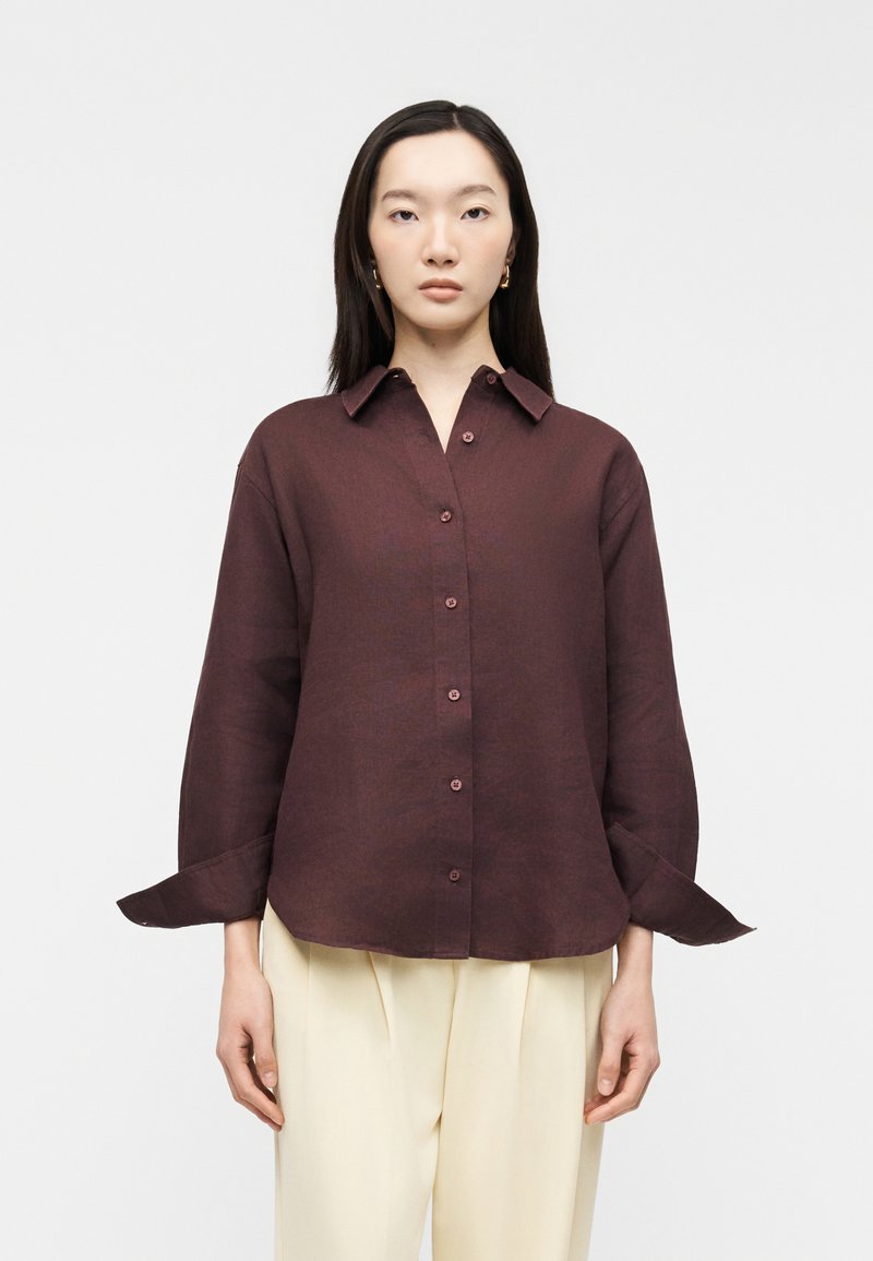 Femme portant une chemise boutonnée marron foncé avec de larges poignets et un pantalon large couleur crème, debout devant un fond blanc uni.