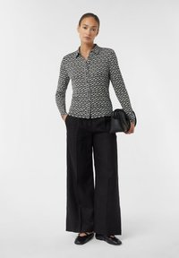 Black and white patterned shirt with a collared neckline, button-up front, and long sleeves, paired with loose black trousers and black shoes.