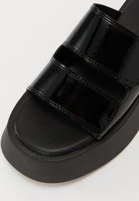Black patent leather slide sandal with two adjustable straps, round toe, and a thick black platform sole. Smooth texture with shiny finish.