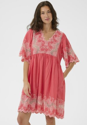 Žena v koralovo ružových šatách s bielou kvetinovou čipkou na rukávoch, výstrihu a leme, stojaca s jednou rukou v vrecku.