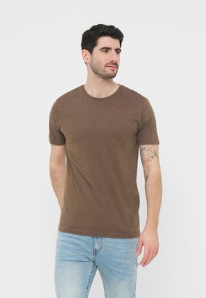 Sötét hajú férfi barna rövid ujjú pólóban és világoskék farmernadrágban, kezét a háta mögött tartva, jobbra néz.