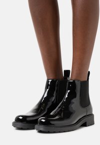Botas Chelsea de charol negro con paneles laterales elásticos, punteras redondeadas y suelas de goma texturizadas. Cuenta con una lengüeta para facilitar su uso.