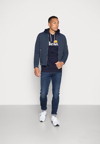 Chaqueta bomber azul marino sobre sudadera azul marino, combinada con jeans azules y zapatillas blancas. La chaqueta cuenta con cierre de cremallera y bolsillos laterales.