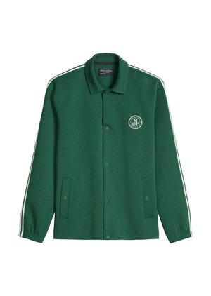 Junger Mann trägt eine grüne Jacke mit Knöpfen, weiße Shorts mit "67"-Aufdruck, weiße Sneaker, hält eine grüne Reisetasche mit dem Logo "Marc O’Polo".