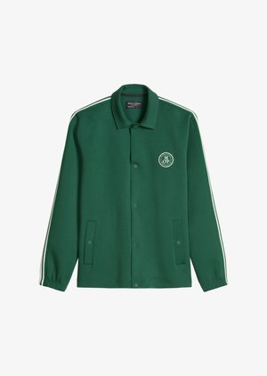 Tânăr purtând o jachetă verde cu nasturi, pantaloni scurți albi cu imprimarea „67”, adidași albi, ținând o geantă verde de sport cu sigla „Marc O’Polo”.